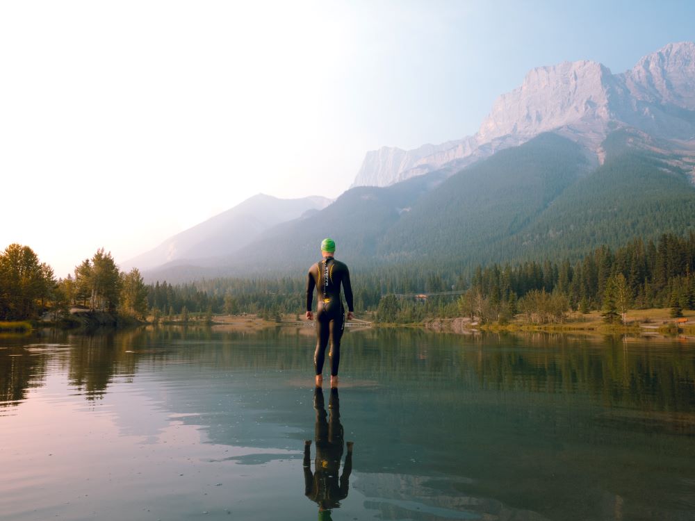 A person in a wetsuit stands in a lake next to a mountain
